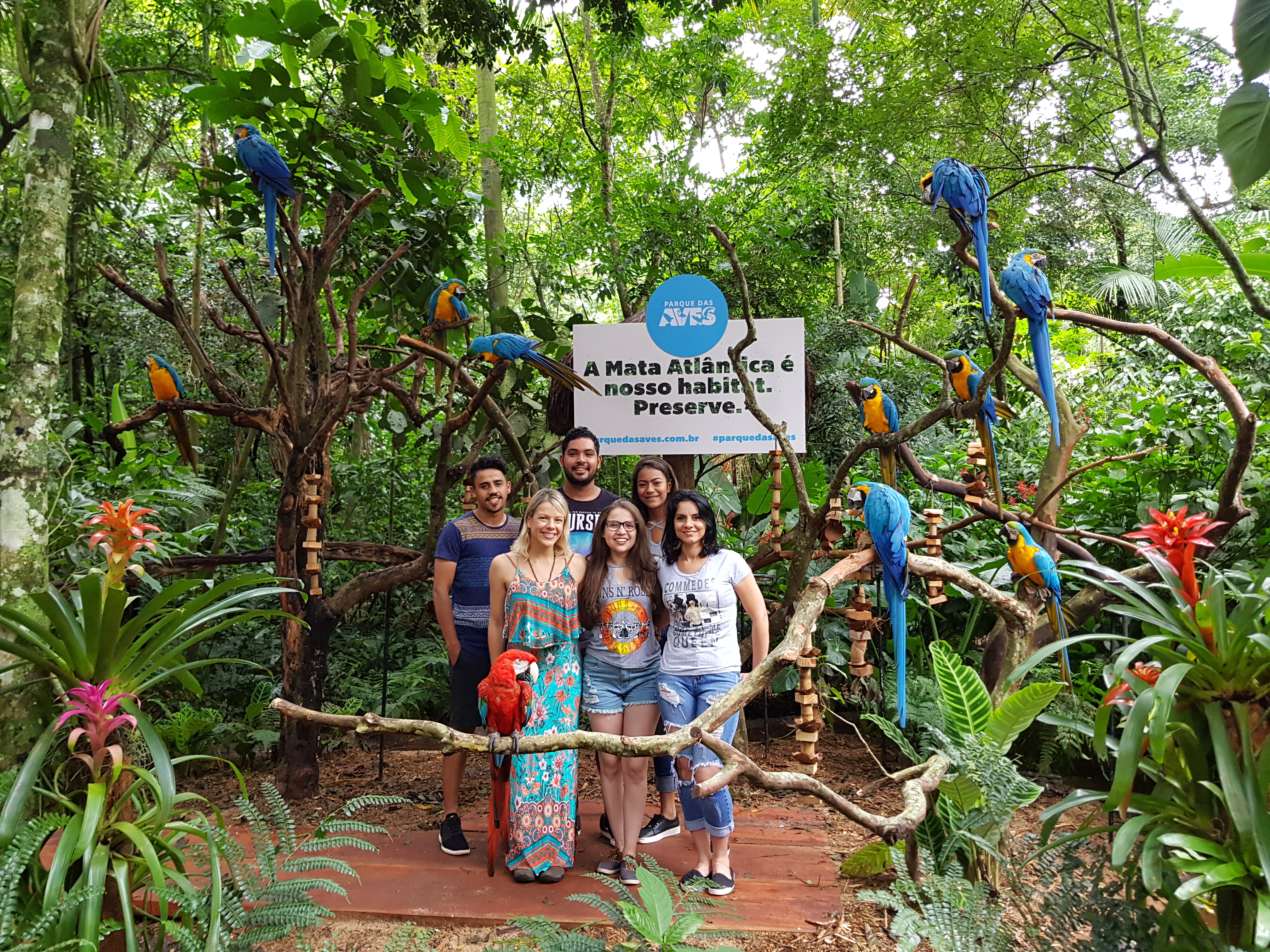 Grupo de seis pessoas sorrindo para a foto no Parque das Aves, cercado por araras coloridas e vegetação da Mata Atlântica, em frente a um painel com a mensagem "A Mata Atlântica é nosso habitat. Preserve."