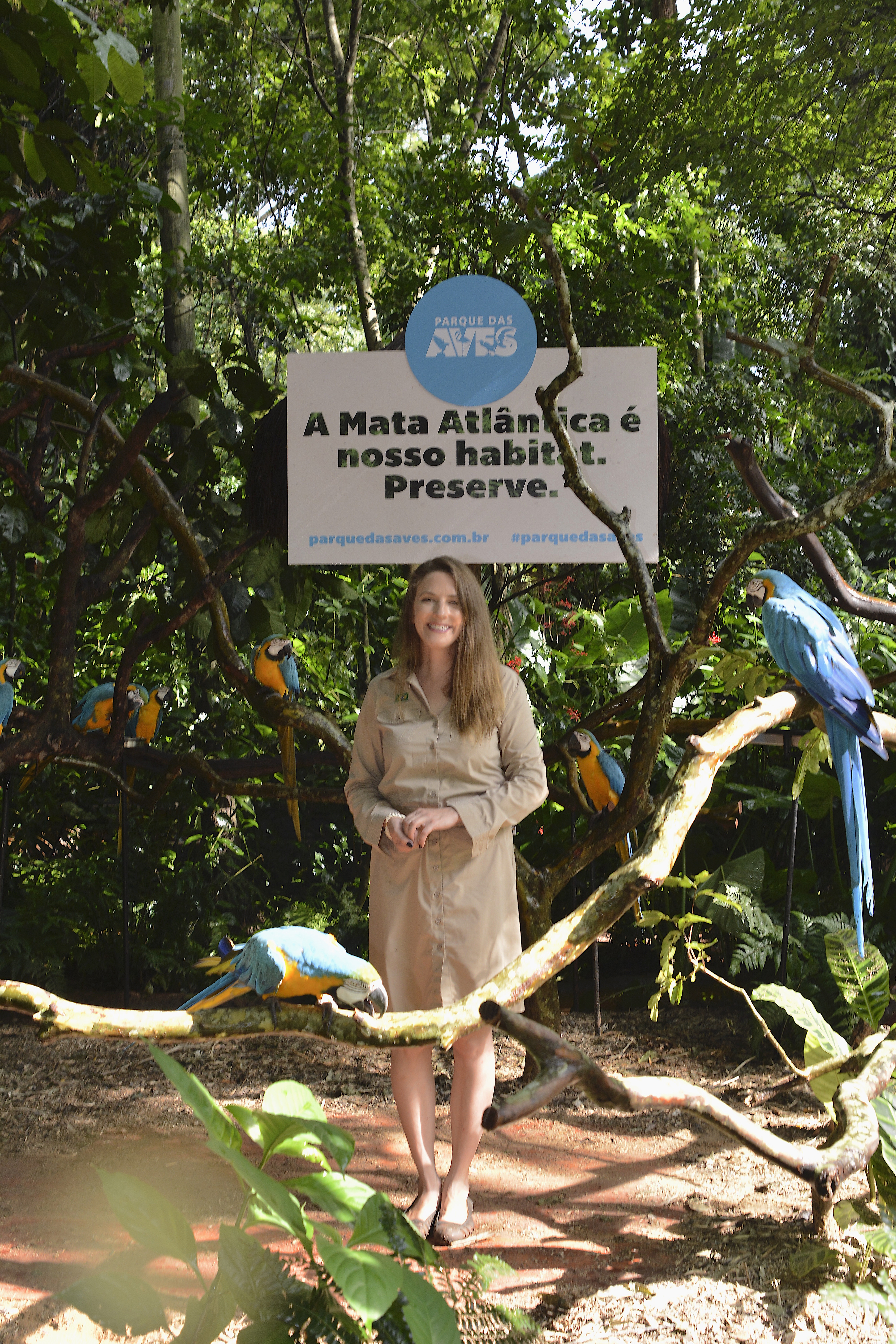Mulher de cabelos longos e loiros, usa uniforme bege do Parque das Aves, posa sorridente em frente a um cartaz que diz "A Mata Atlântica é nosso habitat. Preserve." Araras-canindé (Ara ararauna) estão pousadas nos galhos ao redor, em meio ao vegetação densa da Mata Atlântica.