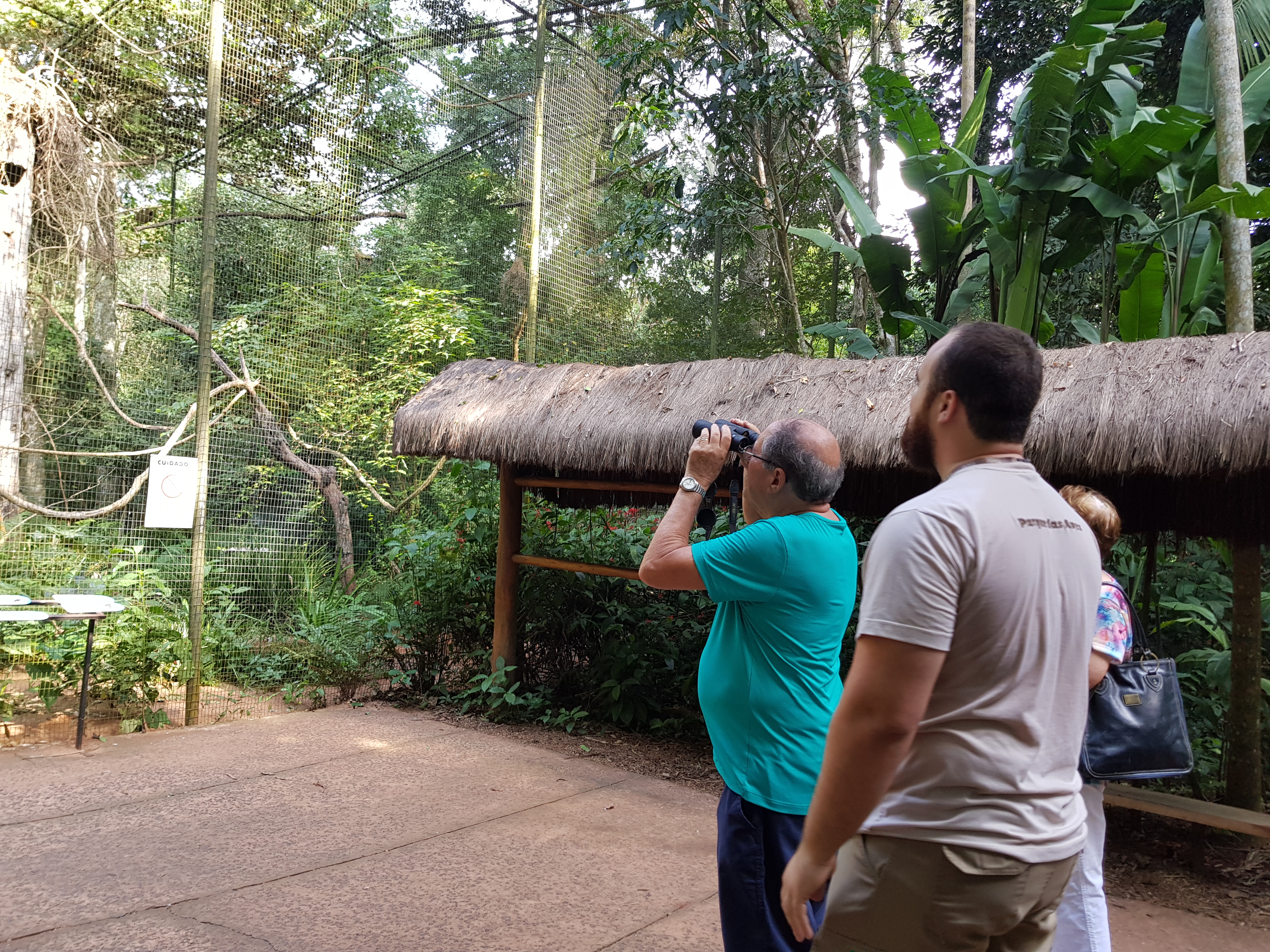  Visitante do Parque das Aves usando binóculos para observar aves dentro de um viveiro, acompanhado por um colaborador do parque.