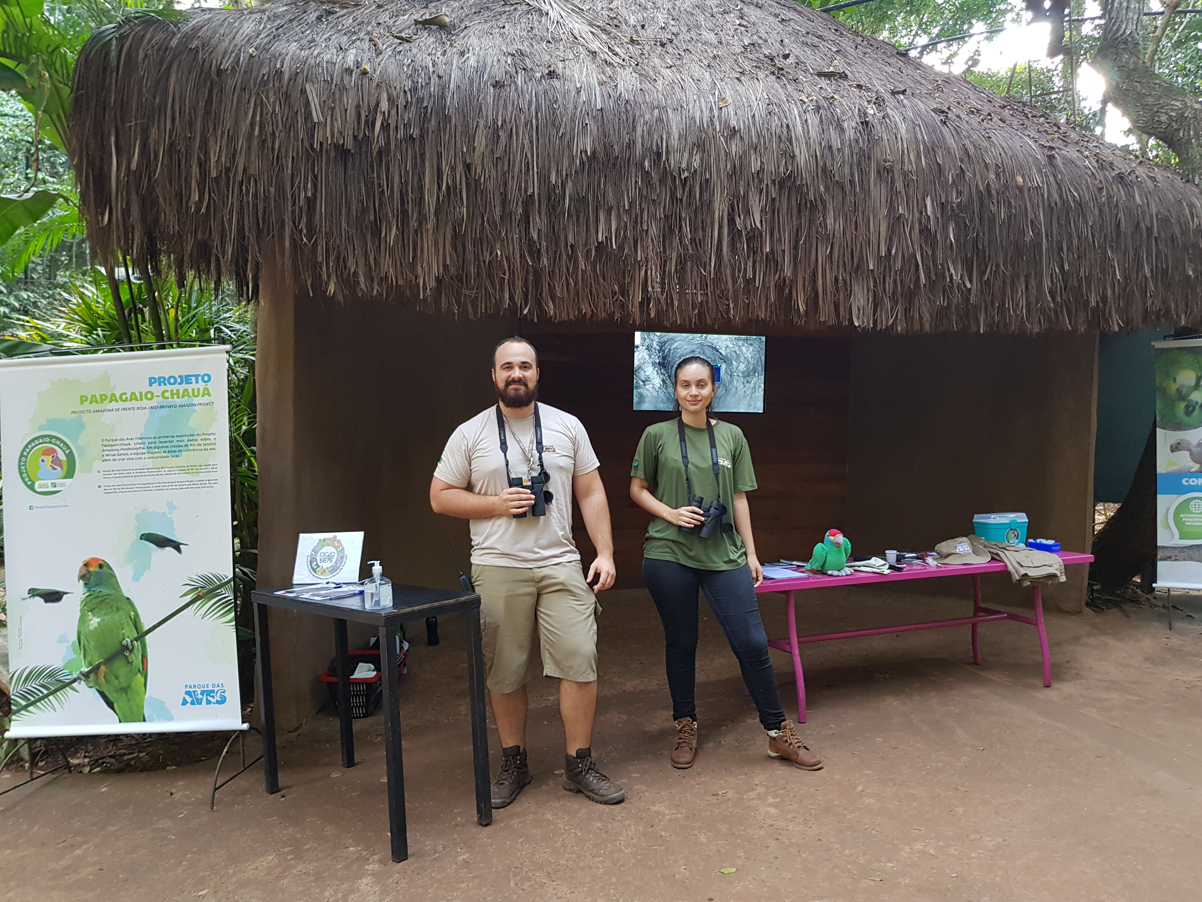 Dois membros da equipe do Parque das Aves, um homem e uma mulher, posam sorrir ao lado de uma mesa com materiais educativos sobre conservação, em frente a uma cabana de palha no Parque das Aves.