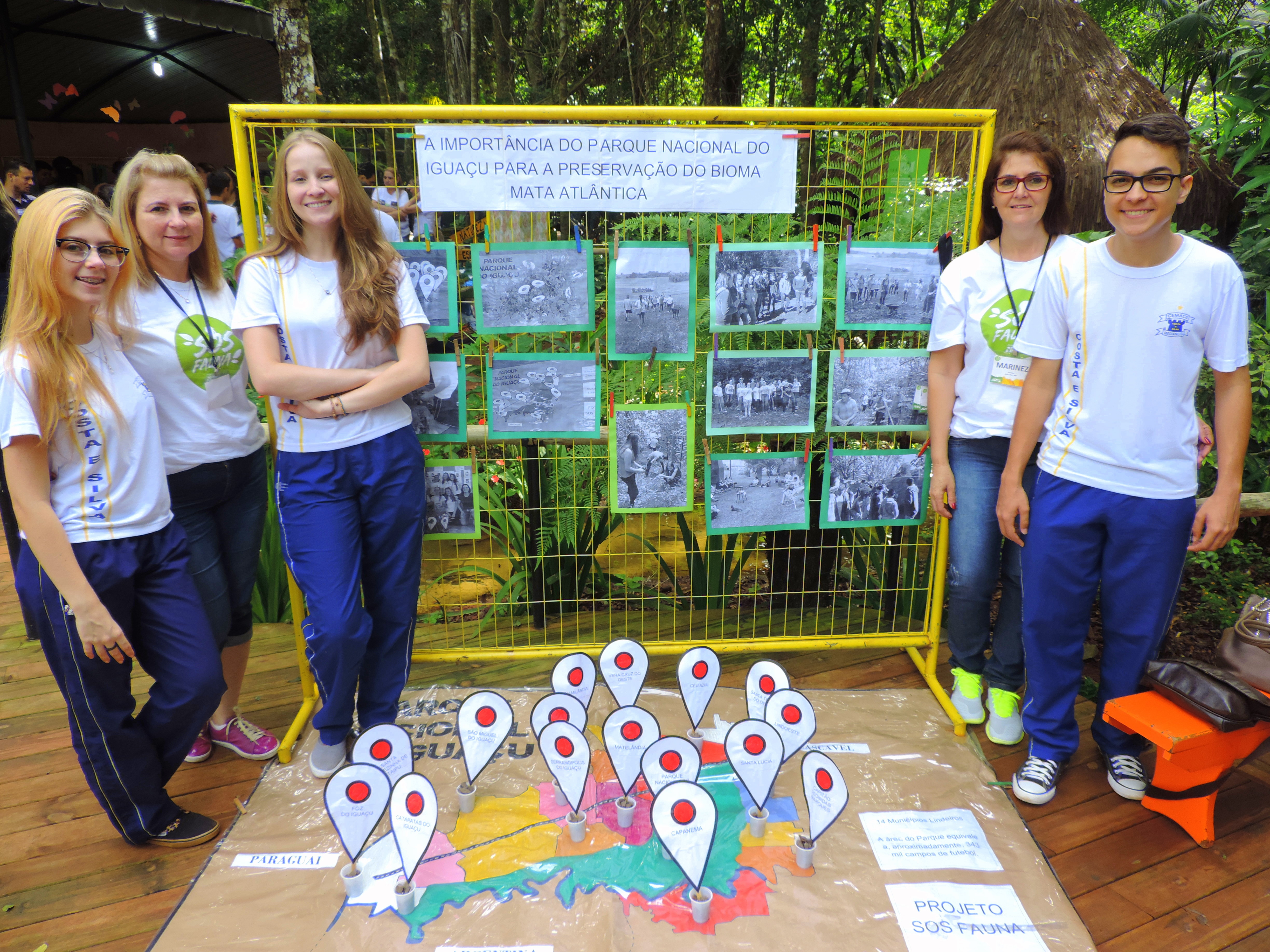 Um grupo de cinco estudantes está posando em frente a um painel informativo sobre a importância do Parque Nacional do Iguaçu para a preservação da Mata Atlântica. O painel exibe várias fotos e informações sobre o projeto. No chão, há um mapa com marcadores indicando locais importantes para o projeto de conservação. Os estudantes estão sorrindo e parecem engajados na apresentação do projeto SOS Fauna.