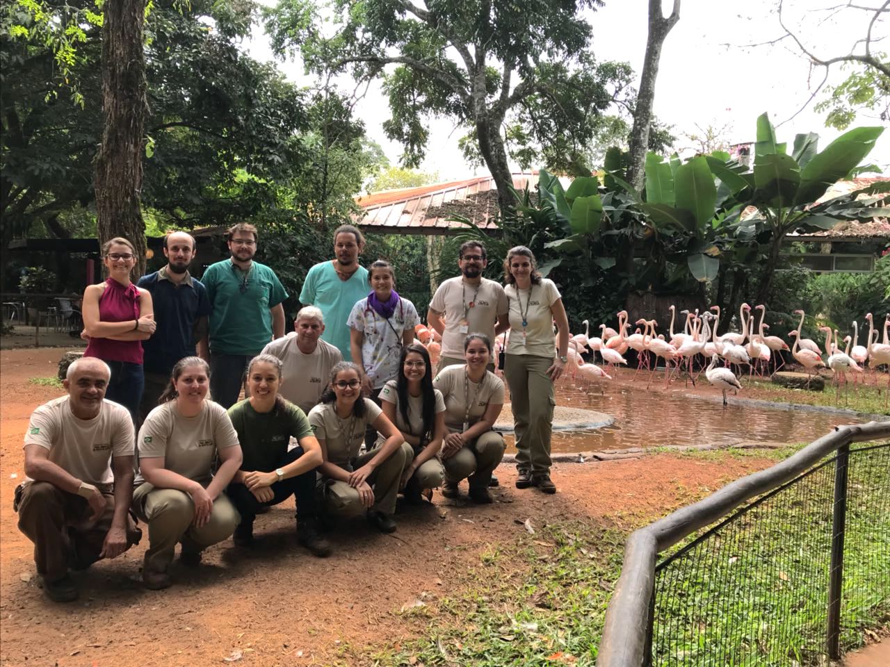 Um grupo de funcionários e pesquisadores está posando para uma foto em frente a um lago com flamingos. As pessoas estão sorrindo e parecem satisfeitas. Ao fundo, vemos uma área arborizada e os flamingos no lago, criando um cenário natural e harmonioso.