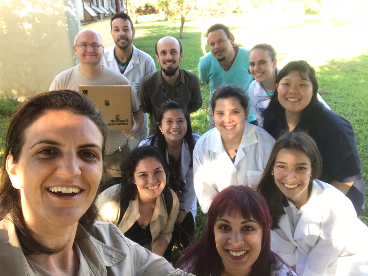Um grupo de pesquisadores está posando para uma selfie ao ar livre, todos sorrindo para a câmera. Alguns estão usando jalecos de laboratório, enquanto outros estão em roupas casuais. Eles estão em um ambiente gramado, sugerindo que estão em um intervalo de suas atividades de pesquisa ou participando de um evento ao ar livre.