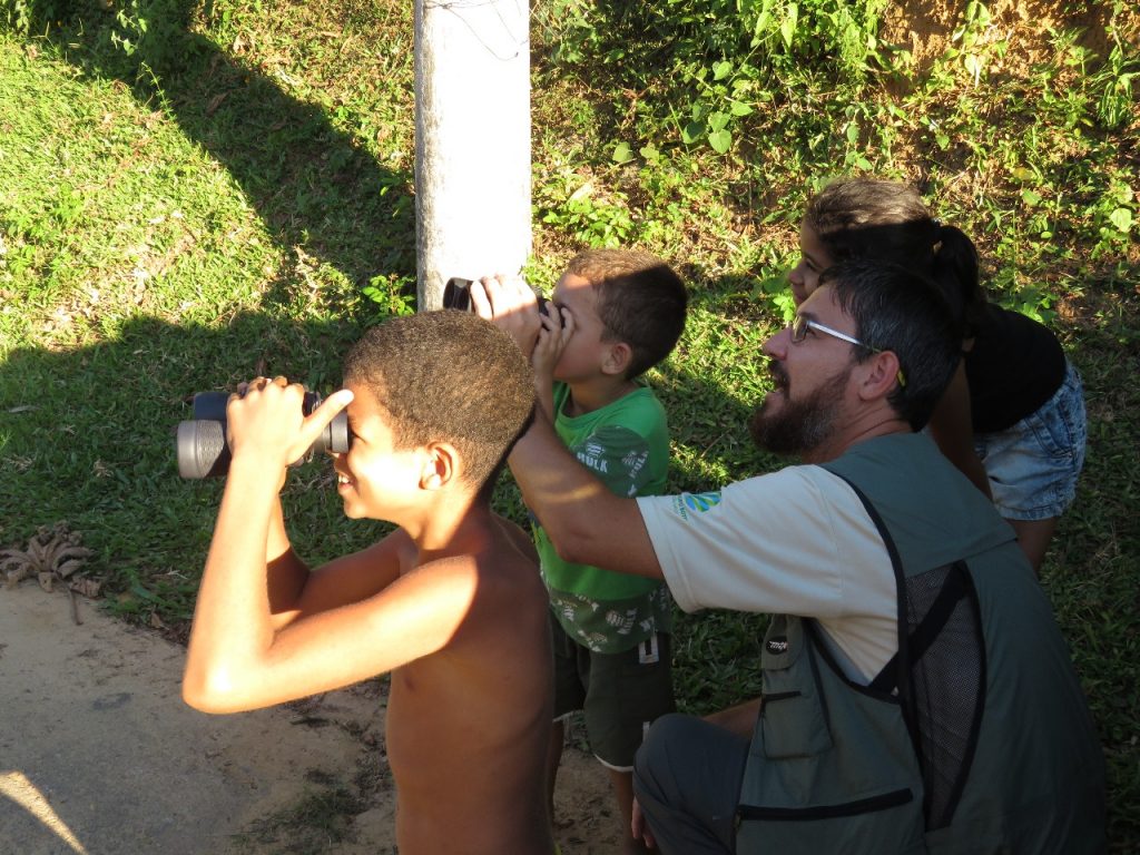 Crianças utilizando binóculos ao lado de um pesquisador em uma área verde.