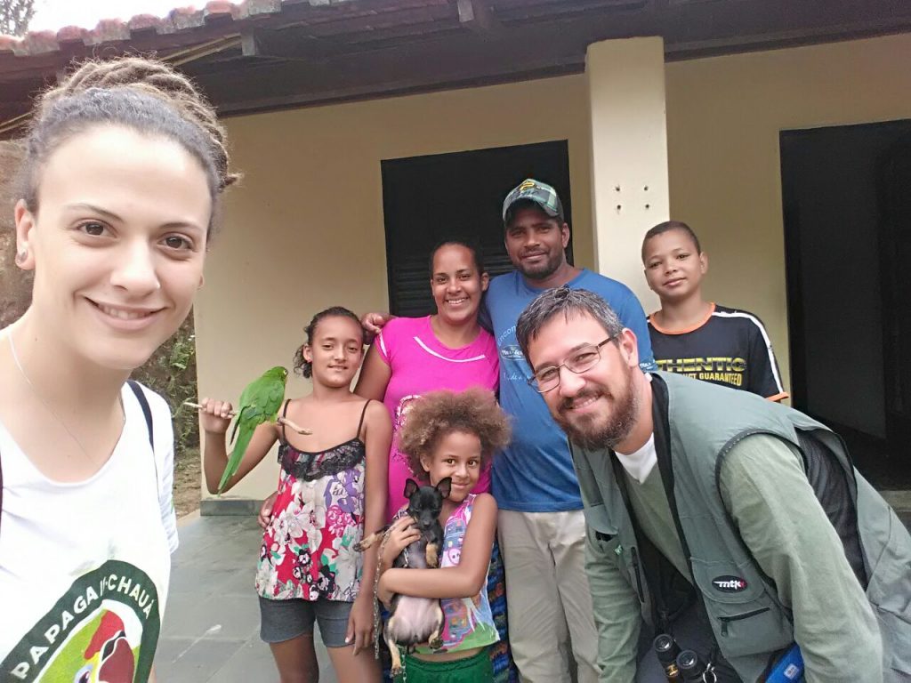 Grupo de pessoas, incluindo pesquisadores e moradores locais, reunidos em frente a uma casa, segurando um papagaio e um cachorro.