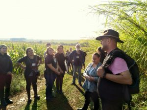 Grupo de pessoas em uma área de plantação ao ar livre, ouvindo atentamente um homem com chapéu e mochila.