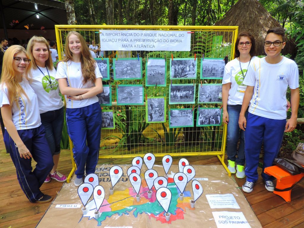Grupo de cinco estudantes ao lado de uma exposição educativa sobre o Parque Nacional do Iguaçu, com fotos históricas e um mapa do parque destacando áreas de preservação da Mata Atlântica.