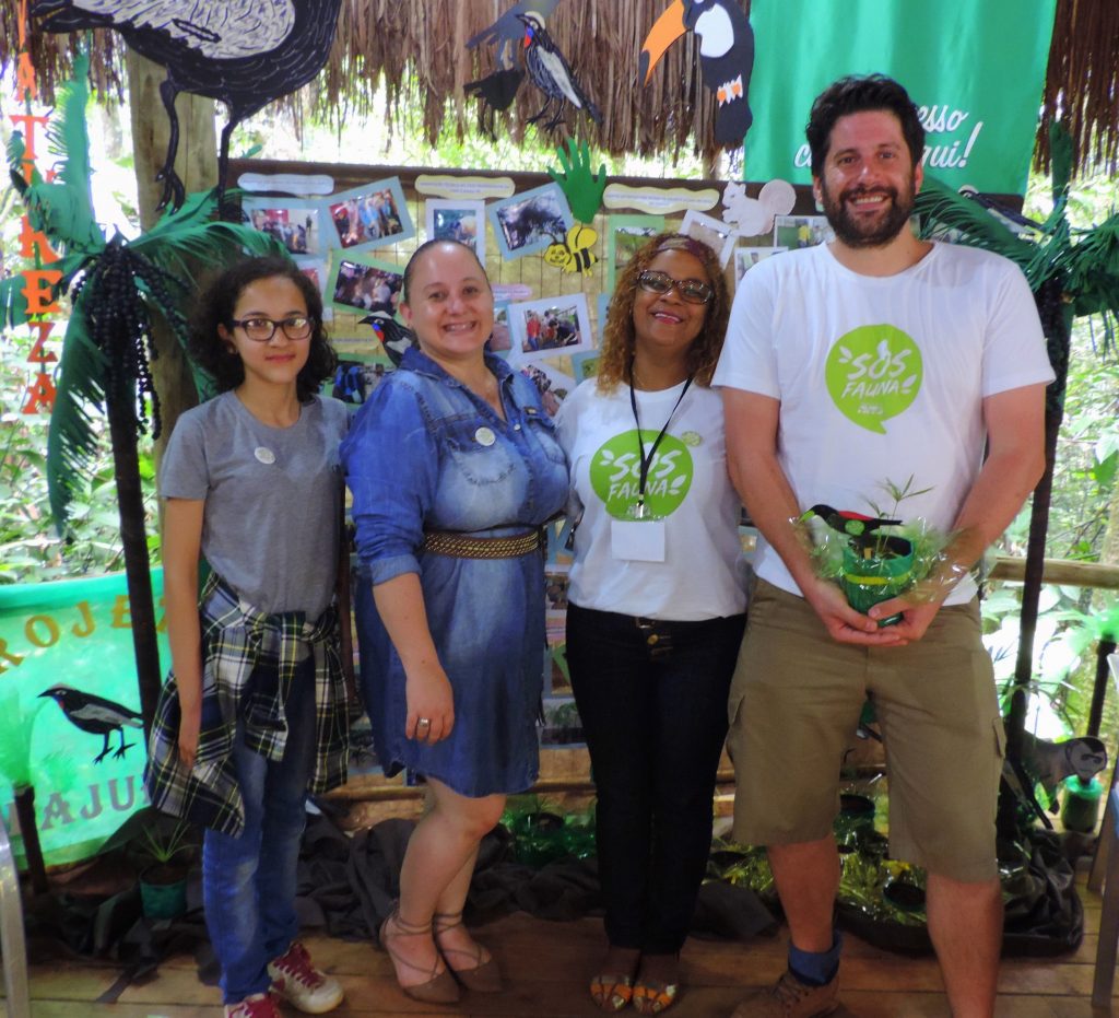 Quatro pessoas posando ao lado de uma exposição educativa sobre o Projeto SOS Fauna, com uma mesa cheia de plantas e representações da fauna local.