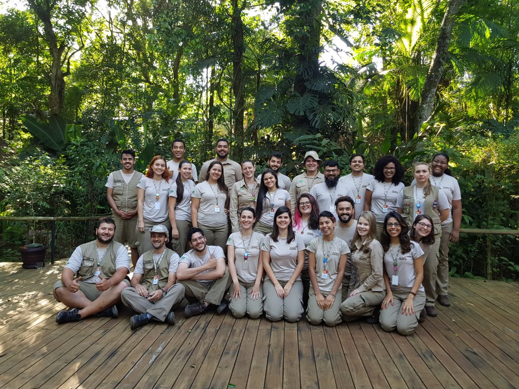 Foto em grupo da equipe do Parque das Aves, composta por cerca de 30 pessoas, todos vestidos com uniformes em toneladas de bege. A equipe está posicionada em uma plataforma de madeira ao ar livre, cercada por uma densa vegetação tropical.  Educação Ambiental
