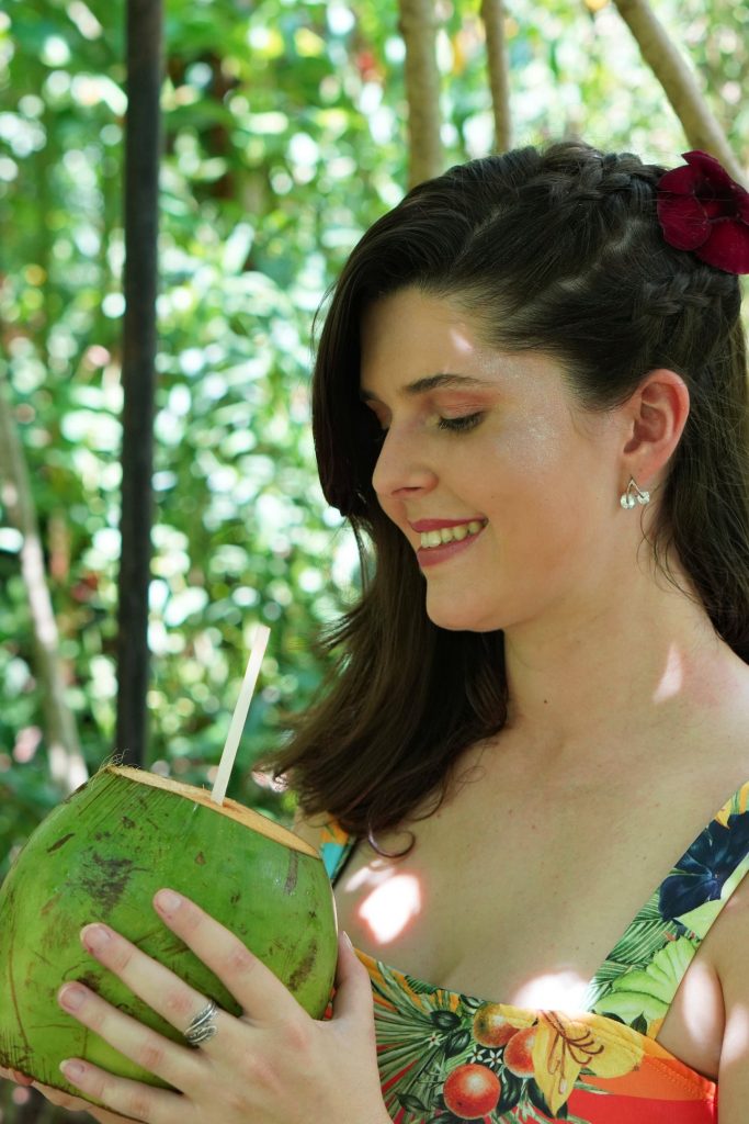 Mulher sorridente segurando um coco verde com um canudo, pronta para beber sua água. Ela veste um vestido colorido e tem flores no cabelo, cercada por um ambiente verde e natural.