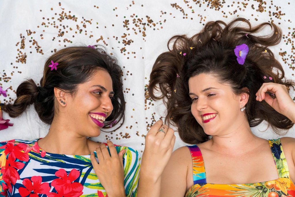 Duas mulheres deitadas lado a lado em uma superfície branca, sorrindo uma para a outra. Elas usam roupas coloridas e têm decorações de flores espalhadas pelo cabelo, criando um visual alegre e festivo.