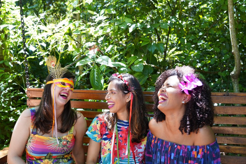 Três mulheres sorridentes sentadas em um banco de madeira em um ambiente ao ar livre, cercadas por vegetação. Elas usam adereços de Carnaval coloridos, como uma máscara com penas, fitas no cabelo e uma flor rosa no cabelo, trazendo um clima festivo à cena.