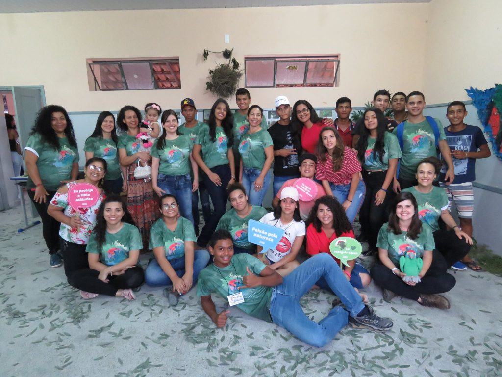 Um grupo de jovens e adultos posa para uma foto em um ambiente interno. Muitos estão usando camisetas verdes com estampa de uma campanha ou evento, segurando placas coloridas com frases e mensagens.