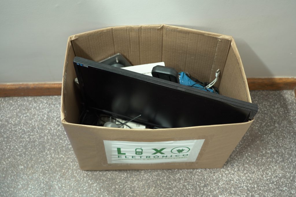 Caixa de papelão com rótulo indicando "Lixo Eletrônico", contendo vários itens eletrônicos descartados, incluindo uma tela e cabos.