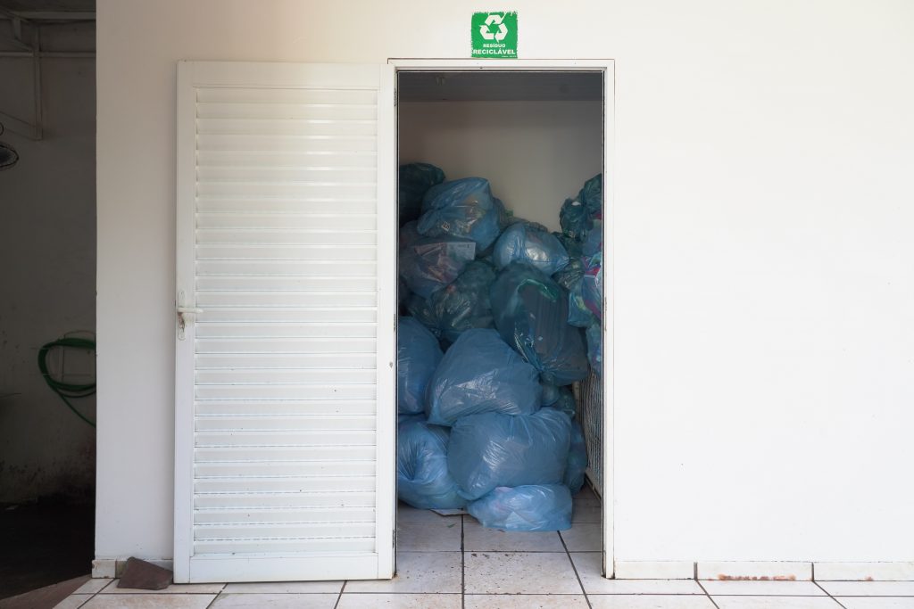 ala com porta aberta, mostrando sacos de lixo azuis empilhados no interior. Acima da porta, há um símbolo de reciclagem verde indicando que o local é destinado à coleta de materiais recicláveis.