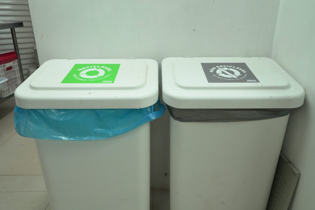 Dois recipientes de lixo com tampas brancas em uma sala. O recipiente à esquerda possui uma etiqueta verde indicando "Orgânico" com um saco de lixo azul, e o recipiente à direita tem uma etiqueta cinza indicando "Não Reciclável" com um saco de lixo preto.