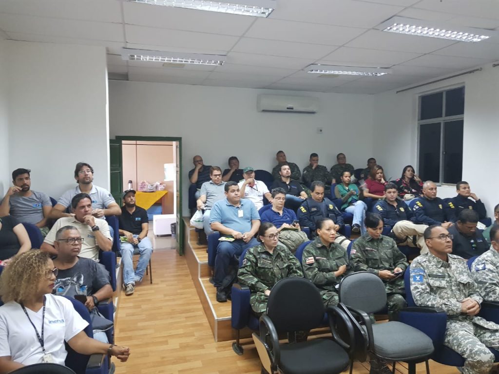 Sala de aula com diversas pessoas sentadas, incluindo membros das forças de segurança em uniformes camuflados e outros profissionais, assistindo a uma apresentação. A sala está equipada com cadeiras em estilo auditório,