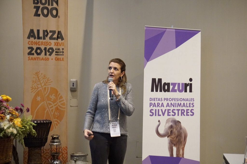 Paloma Bosso, diretora técnica do Parque das Aves, está em pé, segurando um microfone. Em um cartaz próximo a ela está escrito "Mazuri, dieta profesionales para animales silvestres", em espanhol.