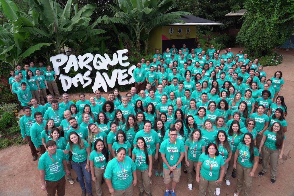 Grande grupo de funcionários do Parque das Aves posando em frente ao letreiro do parque, todos vestindo camisetas verdes.