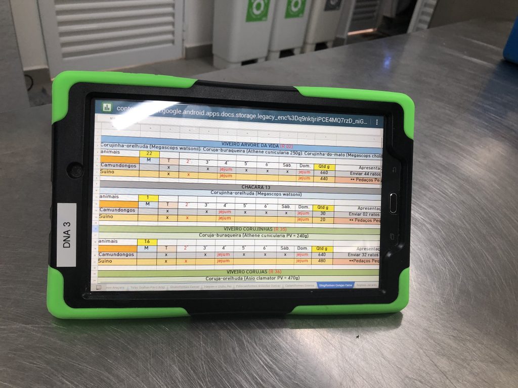 Um tablet com capa protetora verde está sobre uma superfície metálica, exibindo uma planilha com informações coloridas em diferentes colunas. Ao fundo, é possível observar parte de uma bancada e recipientes de armazenamento.