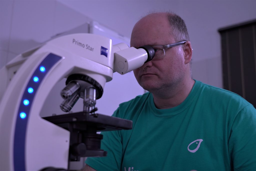 Um homem está sentado e utilizando um microscópio, observando através dos oculares uma amostra.