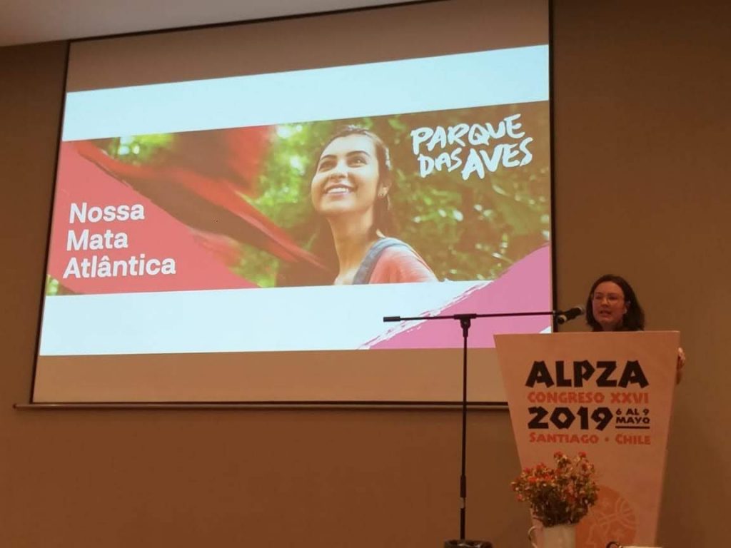 Uma mulher está em um palanque discursando em um congresso, enquanto uma foto é exibida atrás dela uma foto com as frases "Parque das Aves" e "Nossa Mata Atlântica". Na foto uma mulher observa uma arara-vermelha voando.