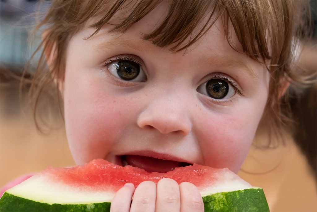 Uma criança de olhos castanhos e cabelo castanho claro morde um pedaço grande de melancia, segurando a fruta com as duas mãos. Seu olhar curioso e a expressão de alegria destacam o momento de prazer ao saborear a fruta.