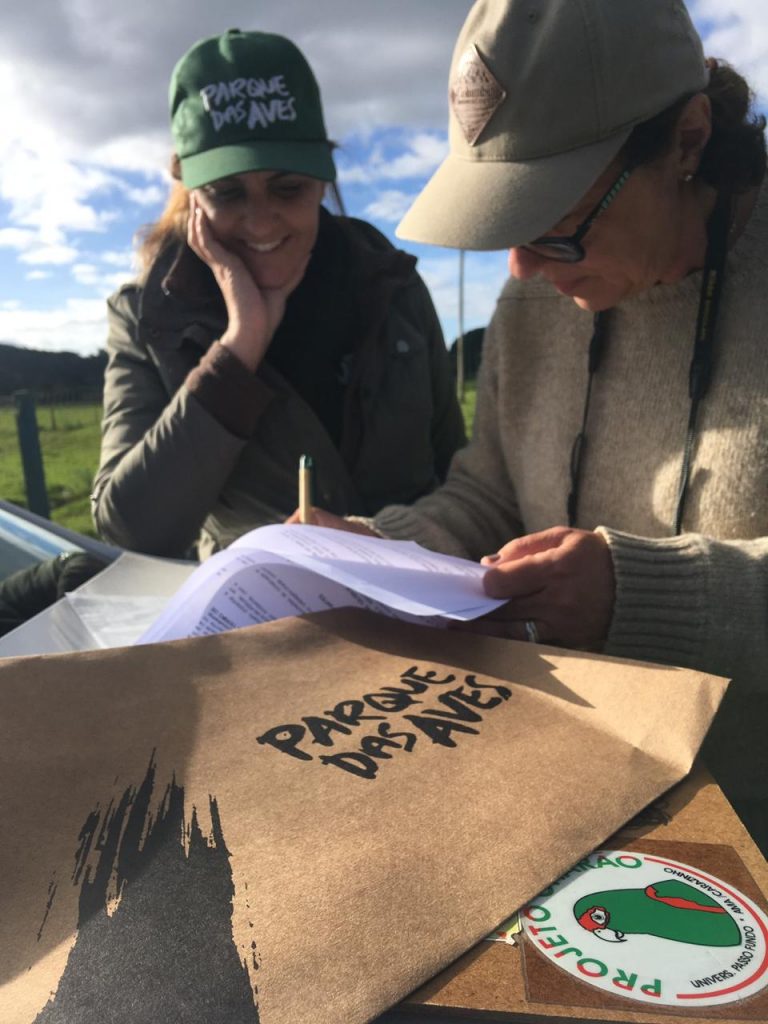 Duas mulheres sentadas próximas. Uma delas está assinando um papel. Vemos também dois envelopes, um está escrito Projeto Charão e em outro lemos Parque das Aves.