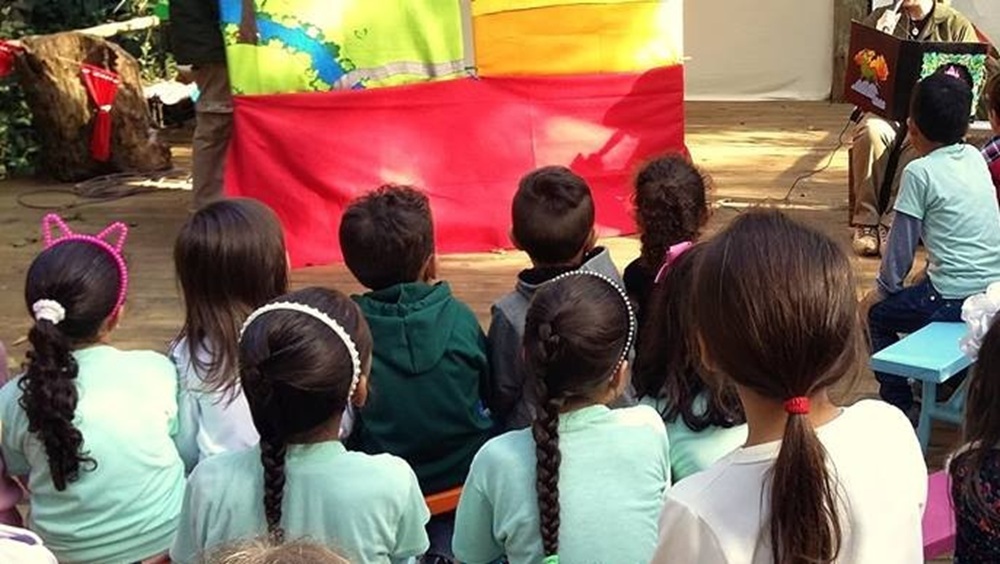 Um grupo de crianças está sentado em frente a um palco ao ar livre, assistindo a uma apresentação sobre educação ambiental. Ao fundo, há um banner com o logotipo do Parque das Aves e a inscrição "Educação Ambiental". A apresentação parece ser lúdica e envolvente, com o objetivo de ensinar as crianças sobre a natureza.