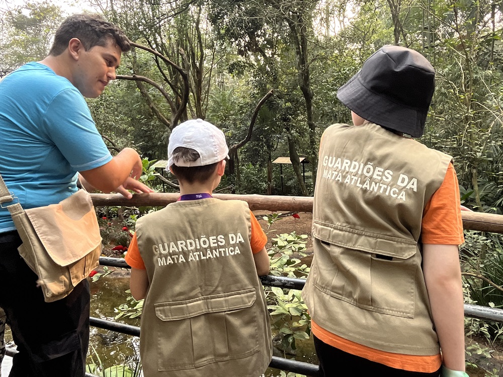 Educador Ambientalconversa com duas crianças usam coletes com a inscrição "GUARDIÕES DA MATA ATLÂNTICA" nas costas. Eles estão em um mirante de madeira, observando a natureza ao redor de um ambiente de floresta.