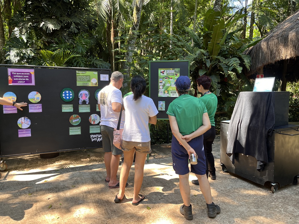 Grupo de visitantes acompanha explicações de educadores diante de painéis informativos sobre colisão de aves com estruturas de vidro.