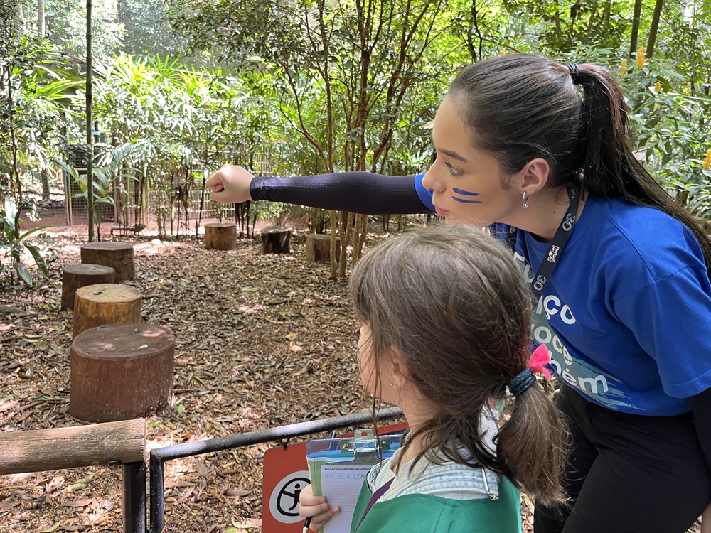 Educadora do Parque das Aves com pintura facial azul aponta algo na trilha enquanto conversa com uma criança segurando uma prancheta de observação.