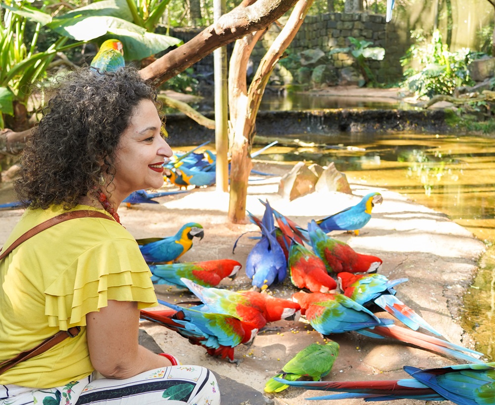 Mulher sorrindo enquanto observa de perto várias araras de cores vibrantes, incluindo vermelho, azul, verde e amarelo, reunidas no chão em um ambiente natural, com um lago e vegetação ao fundo.