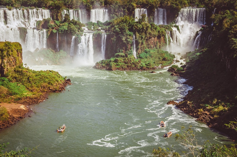 Vista aérea das Cataratas do Iguaçu com várias quedas d'água rodeadas por vegetação densa e verde. No rio, barcos realizam passeios turísticos, proporcionando uma experiência próxima às cataratas