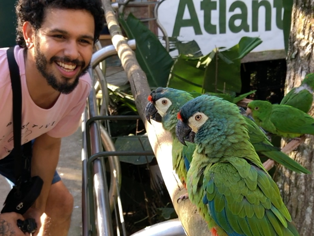 Em um corrimão de madeira vemos duas aves, e no lado esquerdo da imagem vemos uma pessoa sorridente, com barba e abaixada para observar melhor as aves.