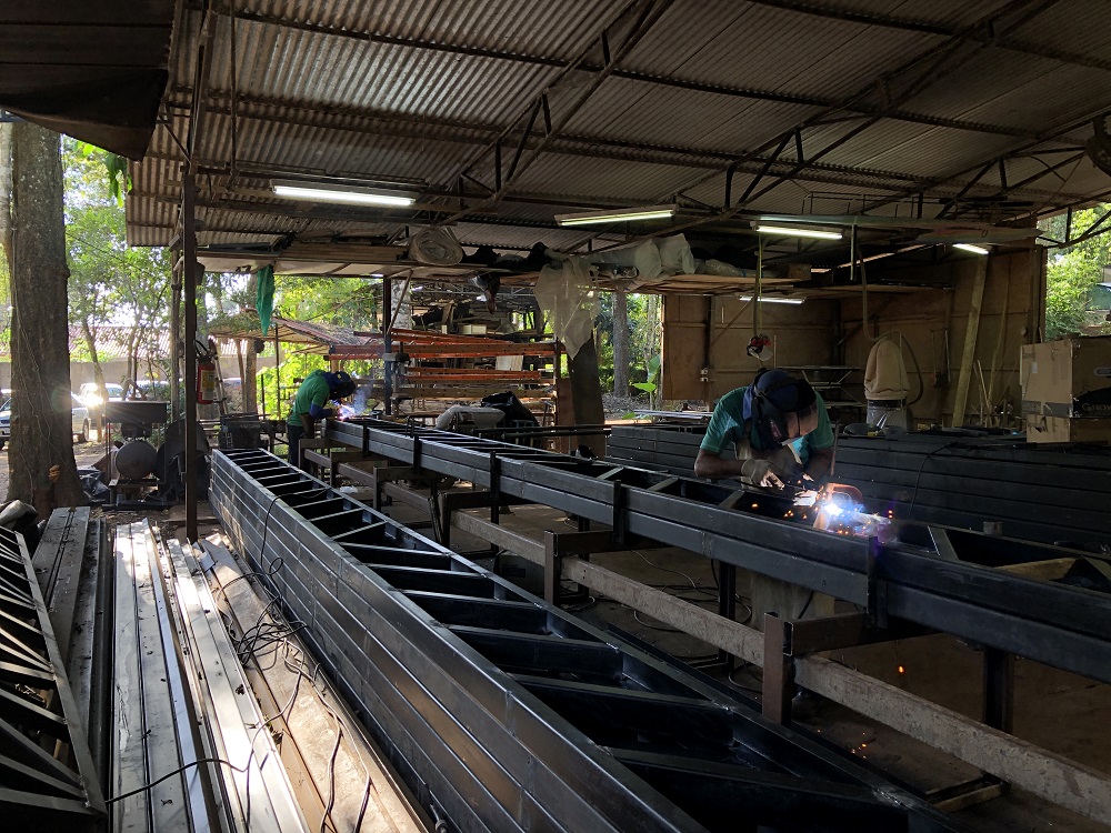 Foto de uma oficina de soldagem onde trabalhadores estão montando treliças metálicas, que são utilizadas como base de sustentação em obras. No primeiro plano, um trabalhador com capacete e luvas de proteção solda uma peça de metal, com faíscas visíveis da soldagem. Ao fundo, outros trabalhadores estão concentrados em suas tarefas, com um foco especial em um soldador que trabalha também soldando a mesma treliça. A cena transmite uma atmosfera de indústria e artesanato, com várias peças de metal e equipamentos de soldagem organizados ao redor.