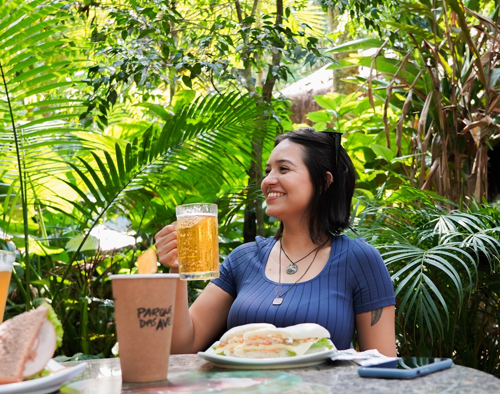 Uma visitante sorridente segurando um copo de cerveja enquanto relaxa em uma mesa ao ar livre cercada pela vegetação exuberante do Parque das Aves. Na mesa, há um sanduíche e um copo personalizado do Parque. No Complexo Gastronômico