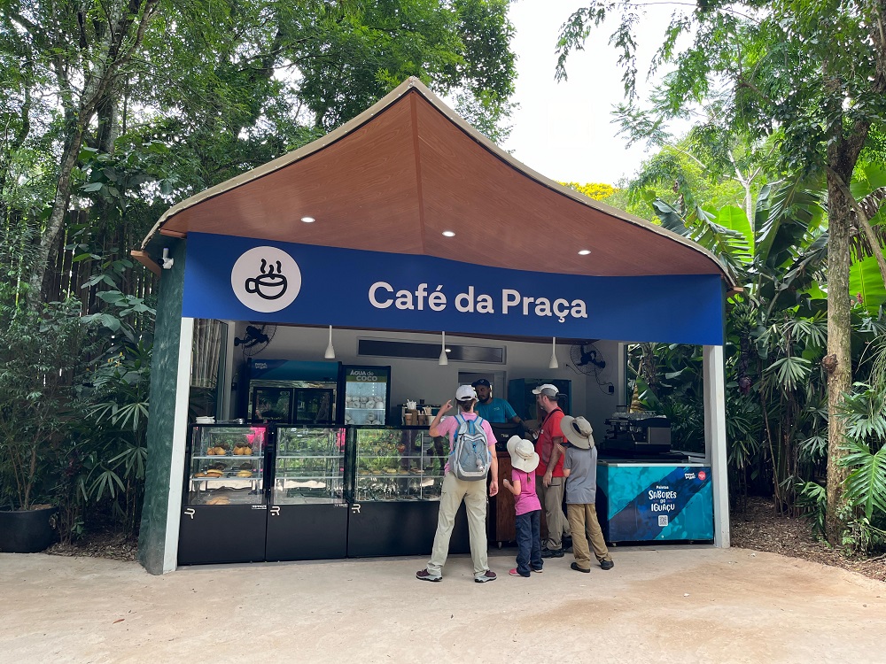 Imagem da fachada do Café da Praça no Parque das Aves. Um grupo de visitantes, incluindo adultos e crianças, está em frente ao balcão do café, pronto para fazer pedidos. A estrutura do café é moderna e rodeada pela vegetação exuberante do parque, proporcionando um ambiente acolhedor para os visitantes, último ponto do Complexo Gastronômico