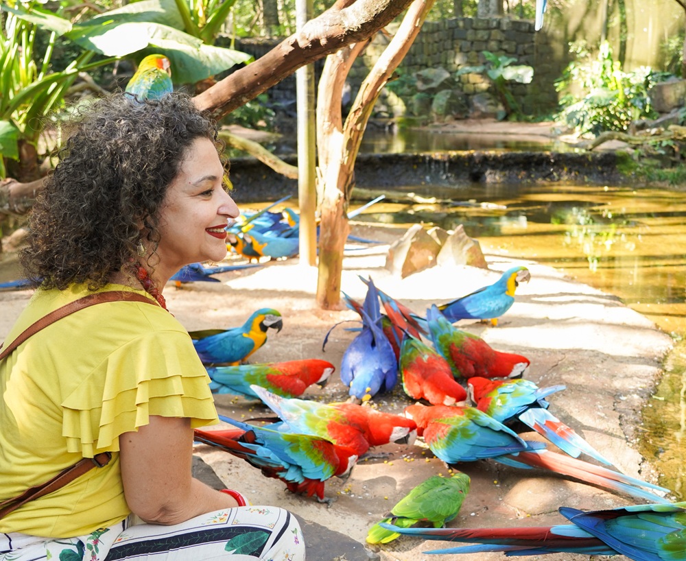 Mulher sentada próxima a um grupo de araras coloridas no chão, em um espaço arborizado e iluminado do Parque das Aves durante o feriado de Carnaval.