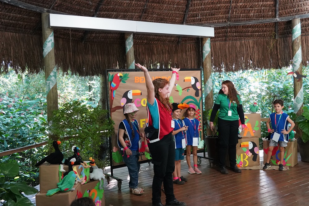Crianças participam de atividades educativas na Colônia de Férias do Parque das Aves, interagindo com educadores em um espaço decorado com elementos da fauna da Mata Atlântica.