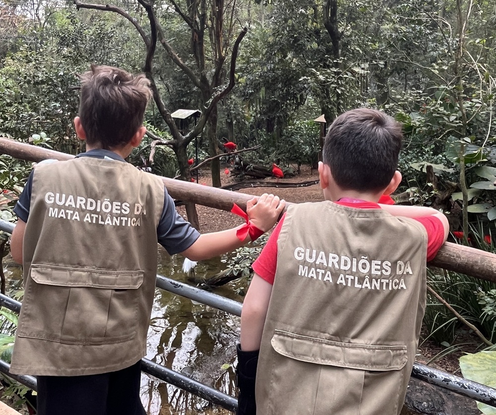 Duas crianças de costas, usando coletes "Guardiões da Mata Atlântica", observando aves em um viveiro no Parque das Aves.