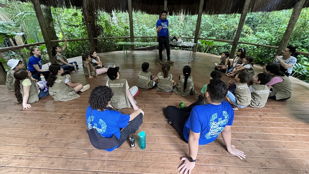Grupo de crianças vestindo coletes "Guardiões da Mata Atlântica" sentadas no chão de um espaço coberto no Parque das Aves, ouvindo atentamente um educador. 