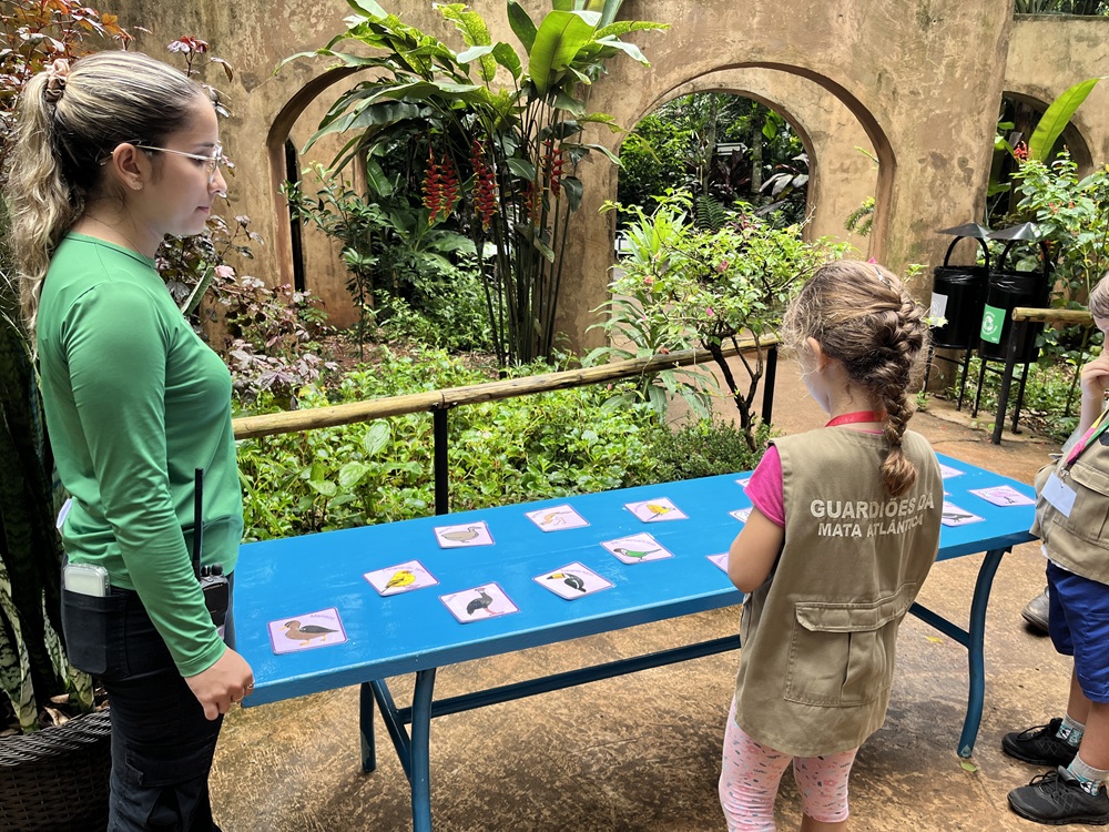 Educadora do Parque das Aves orienta uma criança vestindo o colete "Guardiões da Mata Atlântica" durante uma atividade educativa ao ar livre, com cartas ilustradas de aves sobre uma mesa azul.