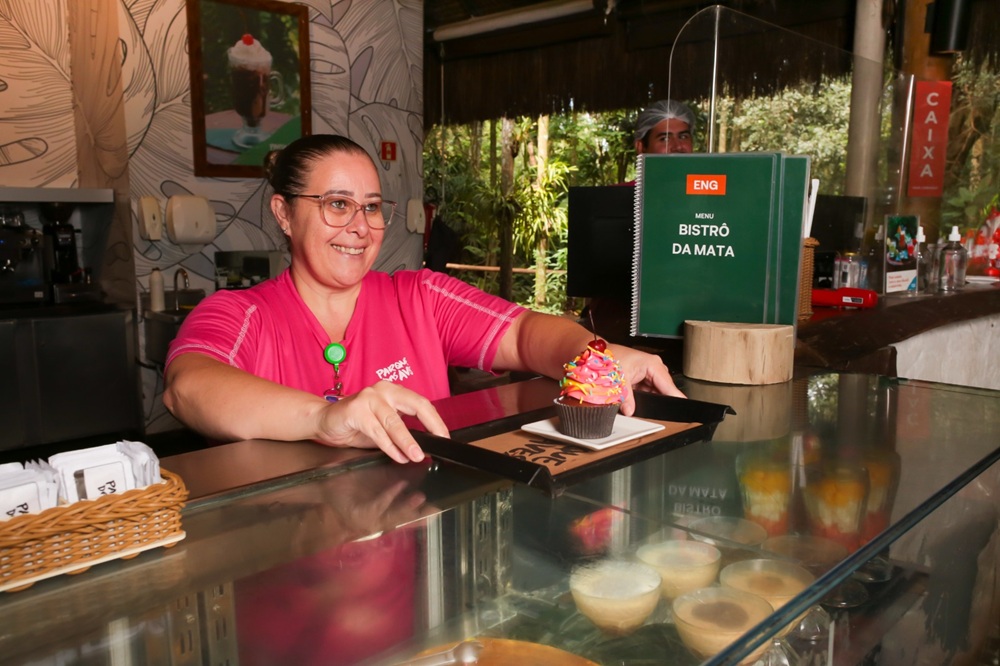 Funcionária do Bistrô da Mata, dentro do Parque das Aves, sorrindo enquanto serve um cupcake colorido sobre um balcão de vidro.