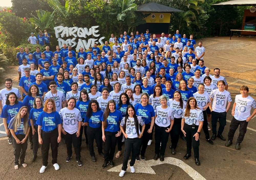 Grande grupo de colaboradores do Parque das Aves reunidos ao ar livre, vestindo camisetas azuis e brancas com a mensagem "Eu faço parte, você também", diante da placa do Parque.