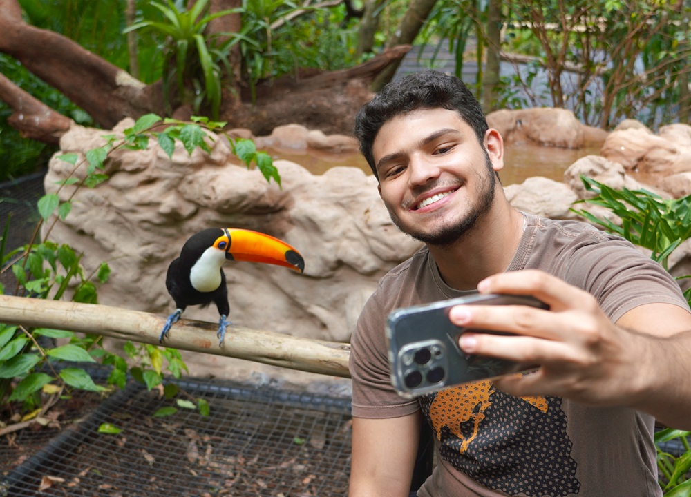 Homem sorridente tirando selfie com um tucano em um viveiro do Parque das Aves.