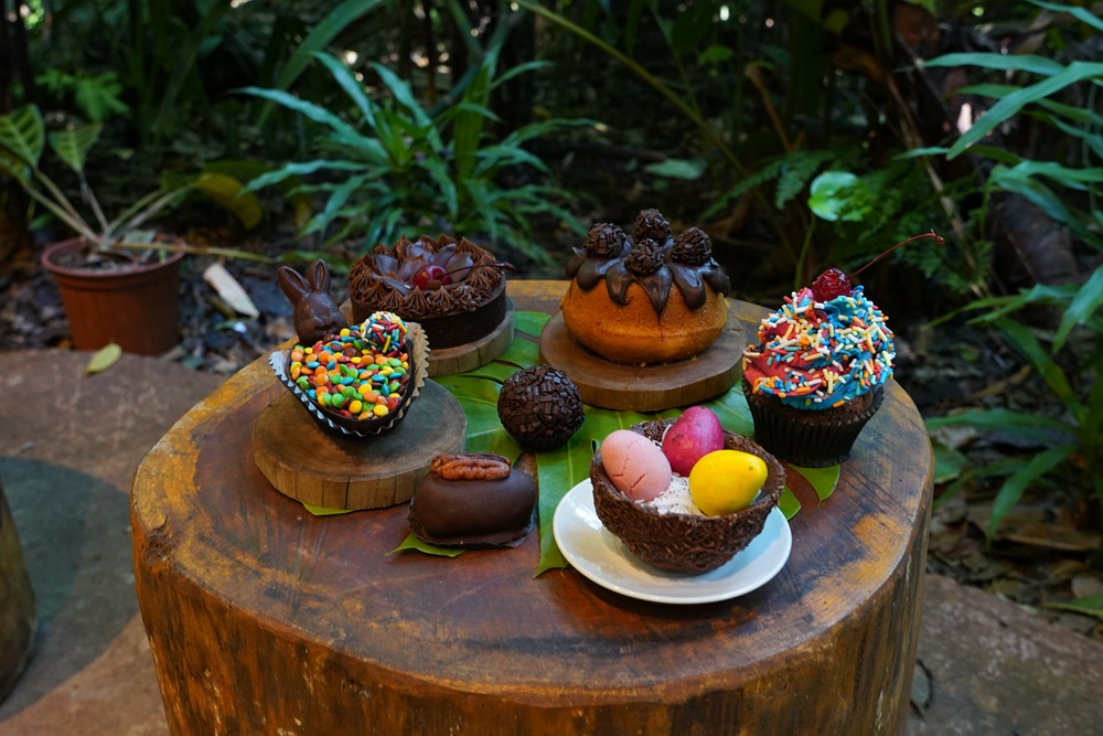 Vários doces temáticos de Páscoa, como cupcakes, trufas, bolos decorados, ovos coloridos e chocolates em formatos variados, estão dispostos sobre um tronco de madeira no meio da vegetação.