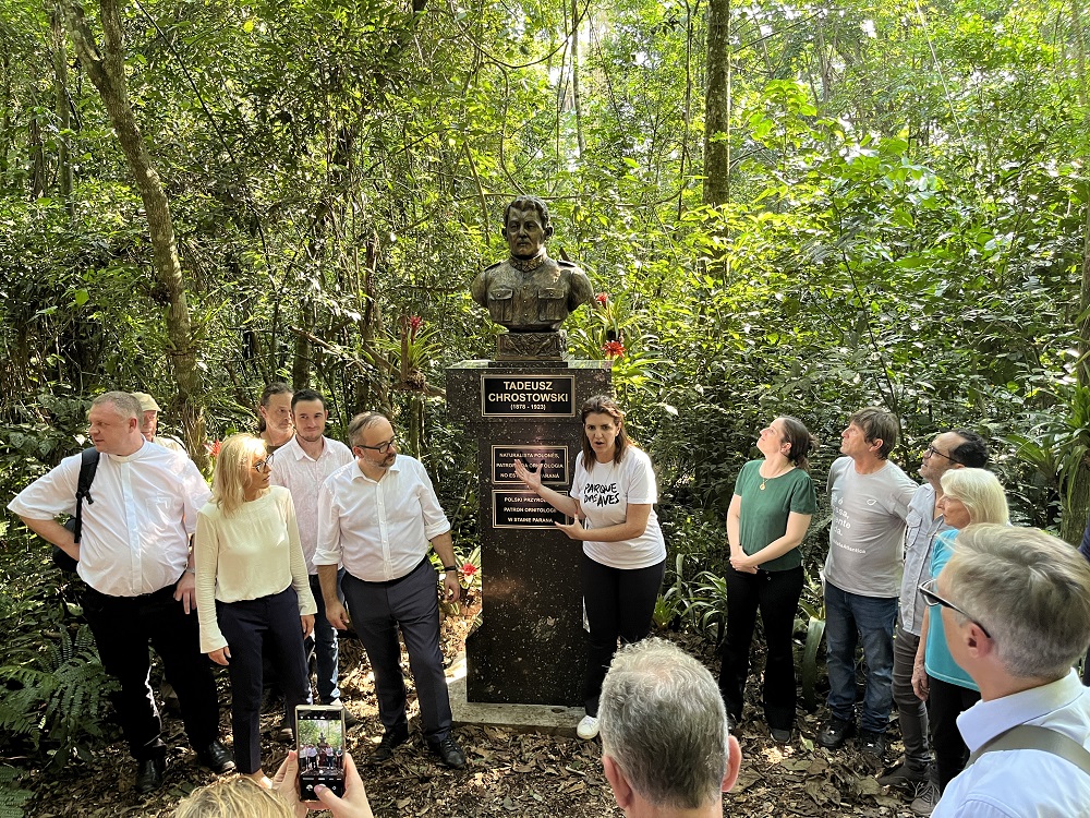  Grupo de pessoas, incluindo representantes da comunidade polonesa, autoridades e colaboradores do Parque das Aves, reunidos ao redor do busto de Tadeusz Chrostowski, instalado na área de floresta do parque.