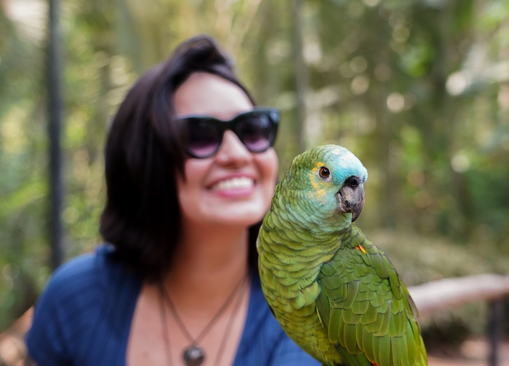 Mulher sorrindo, usando óculos escuros, posa com um papagaio verde pousado em seu braço, em meio à mata do Parque das Aves.