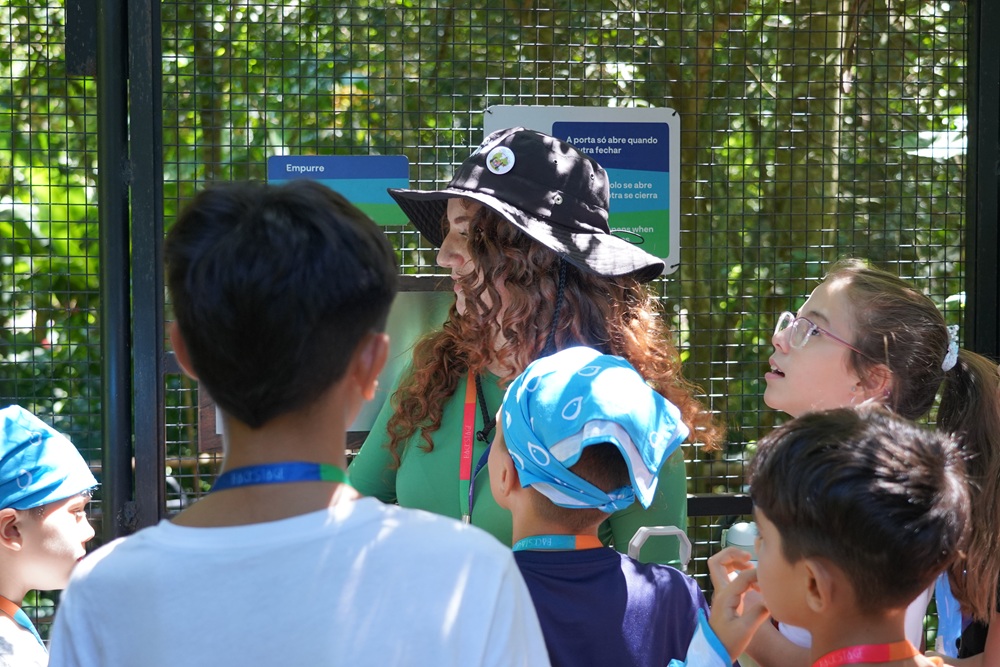 Educadora do Parque das Aves conversa com grupo de crianças na entrada de um viveiro, durante a Colônia de Férias 2024.